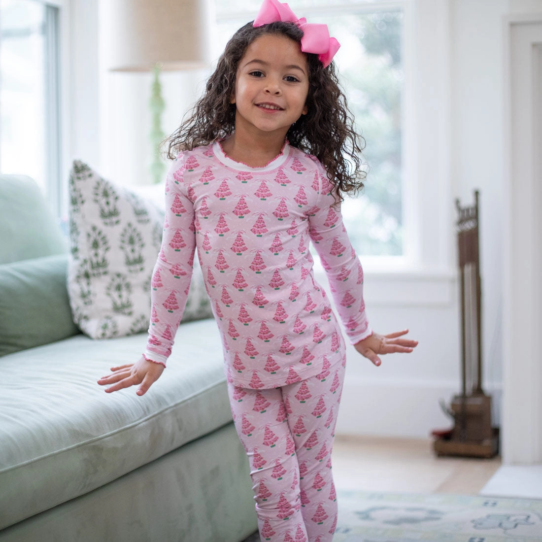 Girl in pink Christmas tree print pajamas standing by a sofa