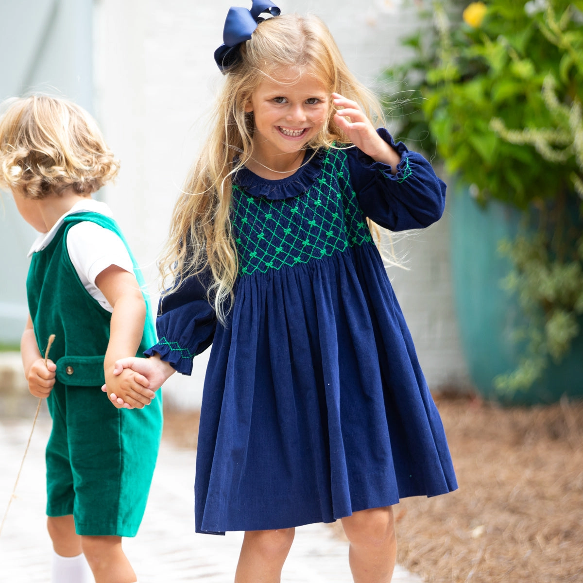 Girl in a navy smocked dress holding hands with a boy in a green romper