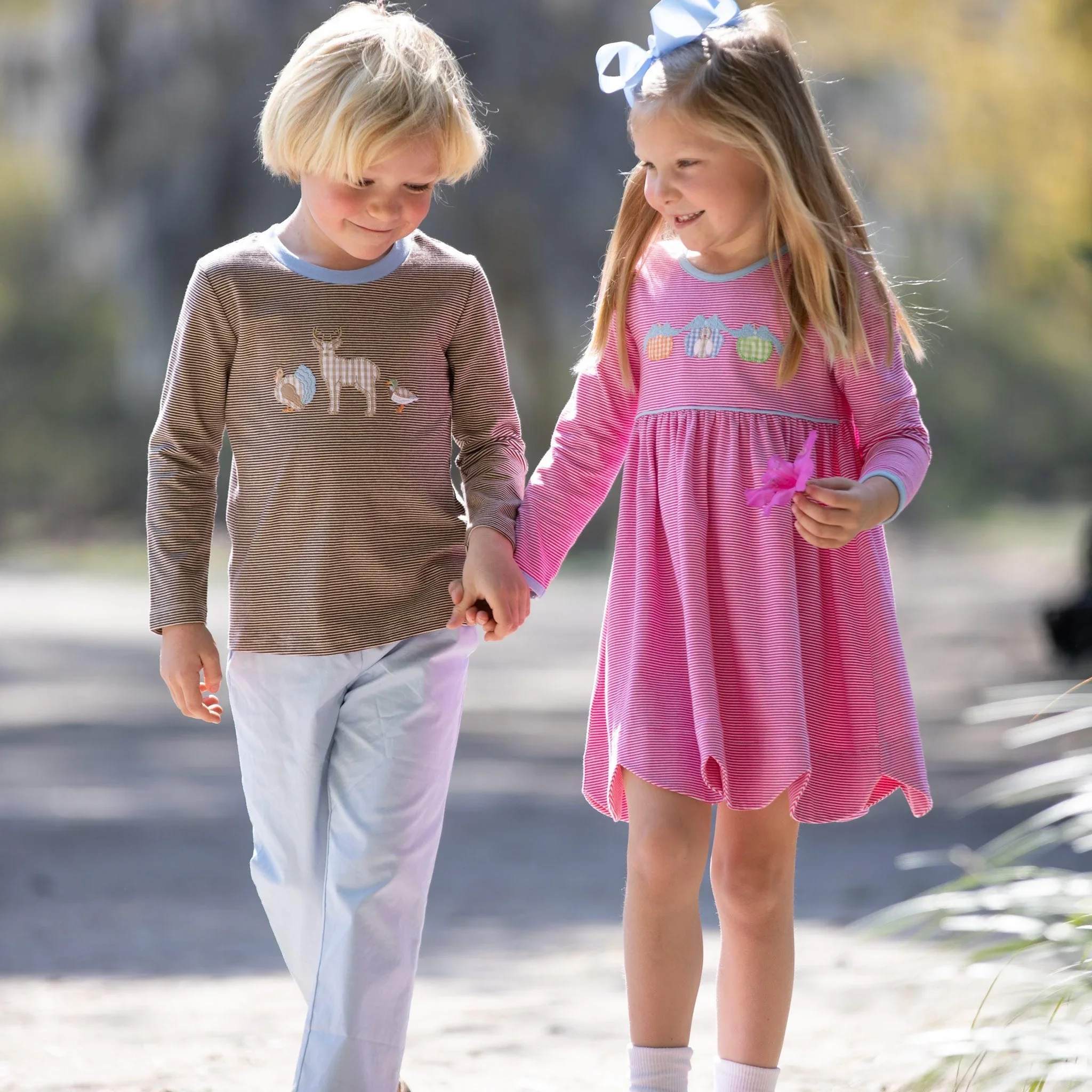 Boy in a brown striped appliqué shirt and girl in a pink striped dress with a blue hair bow, walking hand in hand